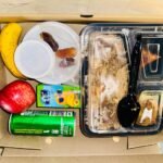 A meal box containing rice, chicken, fruits, drinks, and dates, designed for convenient office lunch catering